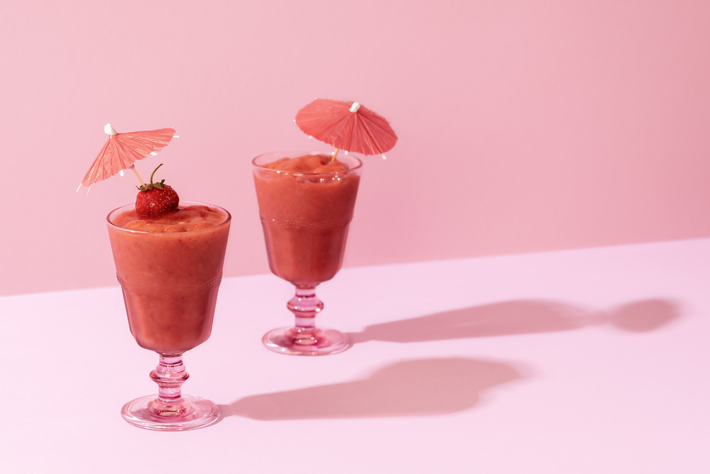 两杯草莓冰沙放在玻璃杯里粉红色背景上有鸡尾酒伞光线刺眼夏季美味的无酒精鸡尾酒健康饮食饮料